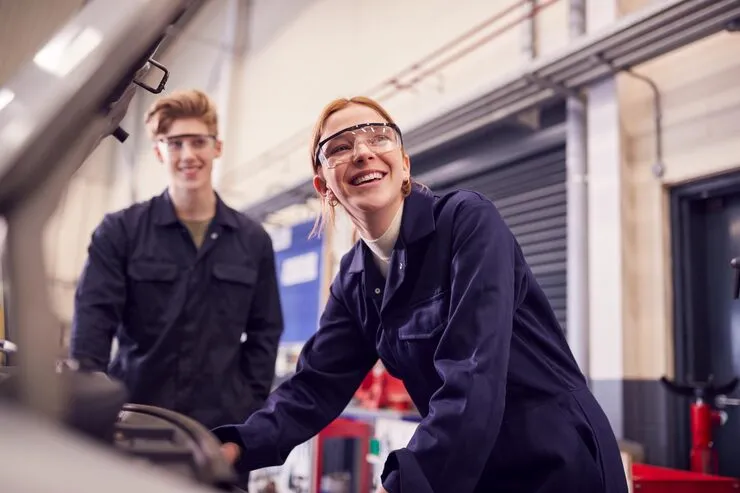 Zwei Auszubildende mit Schutzbrillen lachen bei der Arbeit in einer Werkstatt